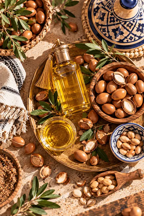 Bottle and bowl of golden argan oil on a brass tray surrounded by argan nuts, kernels and green argan leaves